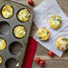Freshly baked egg muffins seasoned with Mccormick Garlic And Herb Pepper Seasoning Ground 4.37 Oz are displayed in a muffin tin and on parchment paper alongside cherry tomatoes.