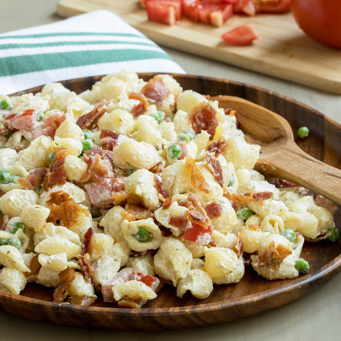 A wooden platter holds a creamy pasta salad featuring shell noodles, peas, and crispy bacon bits seasoned with Mccormick Signature Seasoning Savory Garlic Onion Oz.