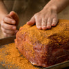 A person is applying a spice rub to a large cut of meat on a baking sheet, preparing it for cooking.