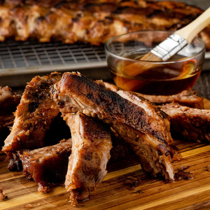 Close-up of juicy, grilled ribs on a wooden cutting board with a bowl of barbecue sauce and a brush.
