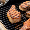 Grilled pork chops with distinct grill marks are cooking on a barbecue grill.