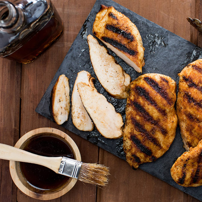 Grilled chicken breasts are arranged on a slate board with a bowl of sauce and a brush, suggesting a barbecue or grilling recipe.
