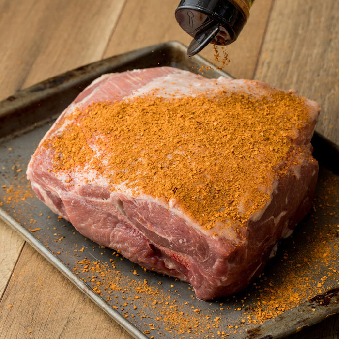A pork shoulder roast is being seasoned with a spice rub on a baking sheet.