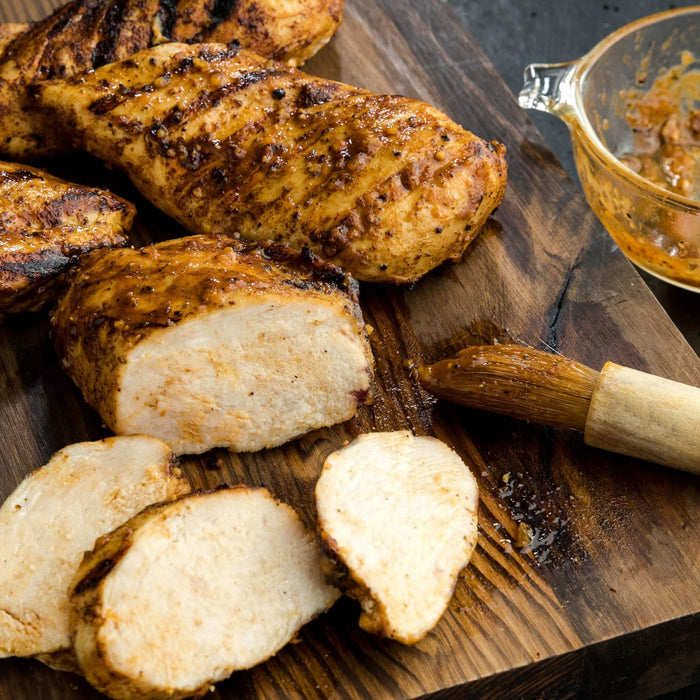 Grilled chicken breasts are sliced and arranged on a wooden board with a brush and a bowl of sauce, showcasing a delicious and healthy meal option.