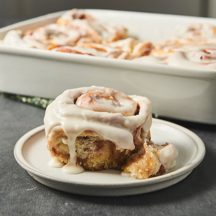 A delicious cinnamon roll with creamy icing sits on a plate with a baking dish full of more rolls in the background.