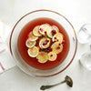 A clear punch bowl filled with a red liquid garnished with lemon and lime slices and pomegranate seeds sits on a table next to a white cloth and glasses with a silver ladle nearby.