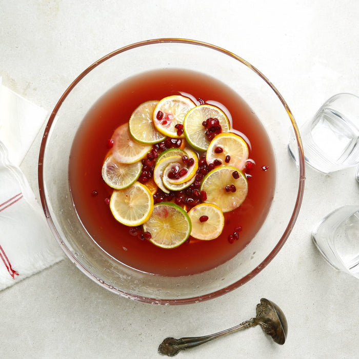 A clear punch bowl filled with a red liquid garnished with lemon and lime slices and pomegranate seeds sits on a table next to a white cloth and glasses with a silver ladle nearby.