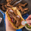 A delicious taco filled with seasoned beef and radish slices is being prepared, with a pan of cooked beef and a bowl of avocado visible in the background.