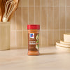 A bottle of Mccormick Perfect Pinch Rotisserie Chicken Seasoning Powder 5 Oz stands on a light marble countertop in a kitchen setting with wooden utensils and stacked plates in the background.