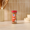 A bottle of Mccormick Perfect Pinch Cajun Seasoning Ground 5 Oz stands on a marble countertop in a bright kitchen setting with cooking utensils and stacked plates in the background.