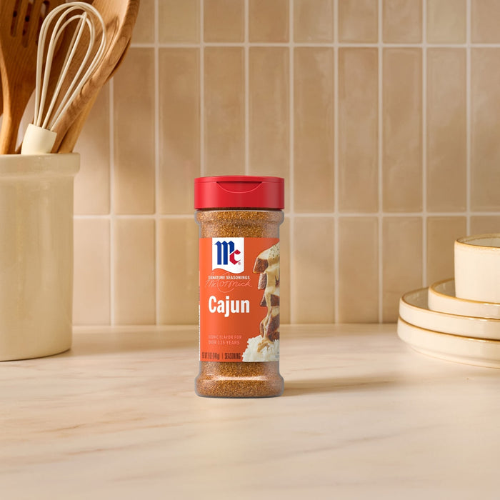 A bottle of Mccormick Perfect Pinch Cajun Seasoning Ground 5 Oz stands on a marble countertop in a bright kitchen setting with cooking utensils and stacked plates in the background.