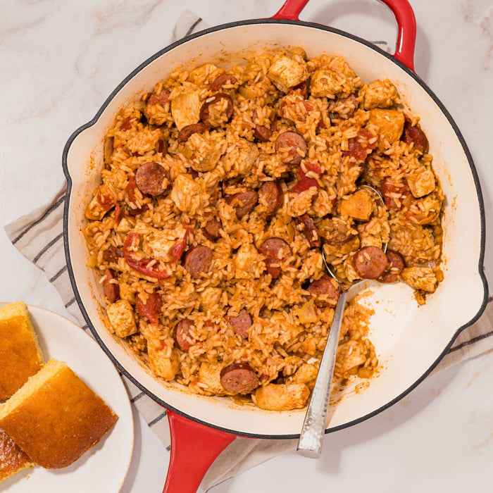 A large red skillet filled with a hearty jambalaya made with rice, chicken, and sausage seasoned with Mccormick Perfect Pinch Cajun Seasoning Ground 5 Oz alongside pieces of golden cornbread.