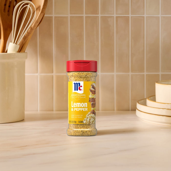 A bottle of Mccormick Signature Seasoning Lemon Pepper Oz stands on a marble countertop in a bright kitchen setting with wooden utensils and stacked plates in the background.
