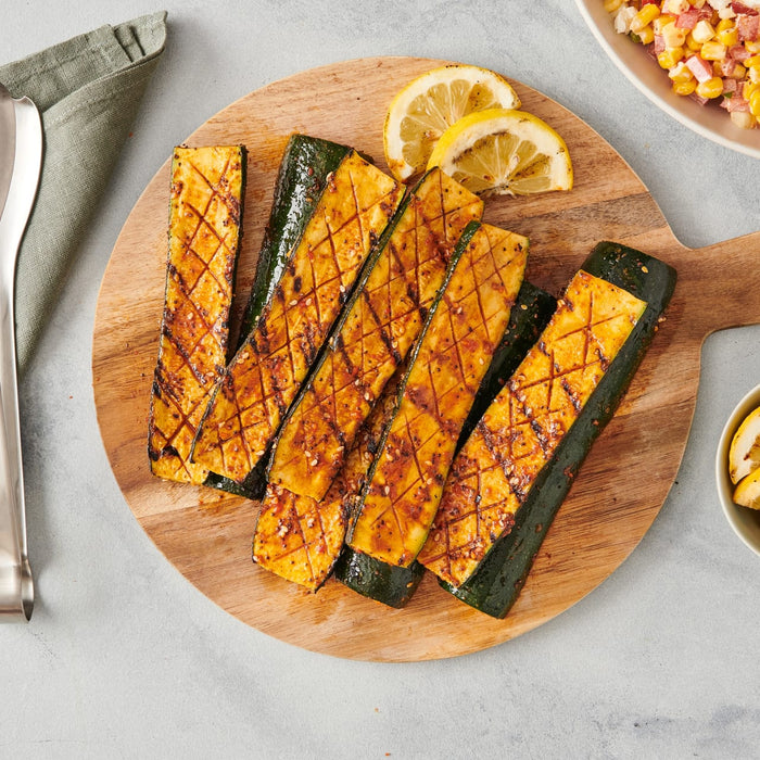 Grilled zucchini spears are seasoned with Mccormick Signature Seasoning Salad Supreme Oz and served on a wooden board with charred lemon slices.