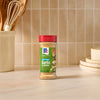 A bottle of Mccormick Garlic And Herb Seasoning Ground 4.37 Oz stands on a light marble countertop in a kitchen setting with wooden utensils and stacked plates in the background.