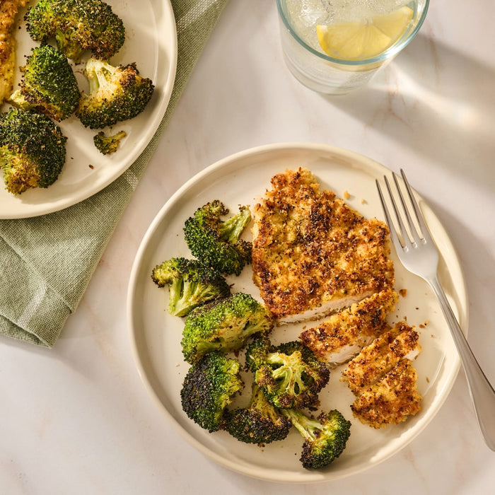 A plate of breaded chicken and roasted broccoli florets seasoned with Mccormick Garlic And Herb Seasoning Ground 4.37 Oz sits next to a glass of lemon water.
