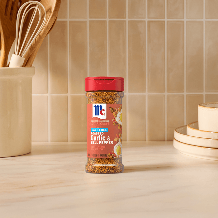 A bottle of Mccormick Roasted Garlic And Red Bell Pepper Seasoning Ground 4.34 Oz stands on a marble countertop in a kitchen setting with wooden utensils and stacked plates in the background.