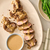 A sliced pork tenderloin is drizzled with a creamy sauce and garnished with Mccormick Onion And Herb Seasoning Crystal 4.16 Oz alongside a bowl of extra sauce and fresh green beans.