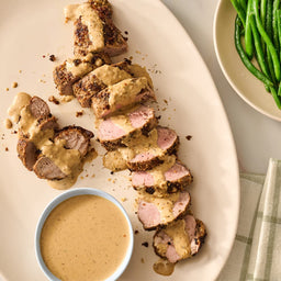 A sliced pork tenderloin is drizzled with a creamy sauce and garnished with Mccormick Onion And Herb Seasoning Crystal 4.16 Oz alongside a bowl of extra sauce and fresh green beans.
