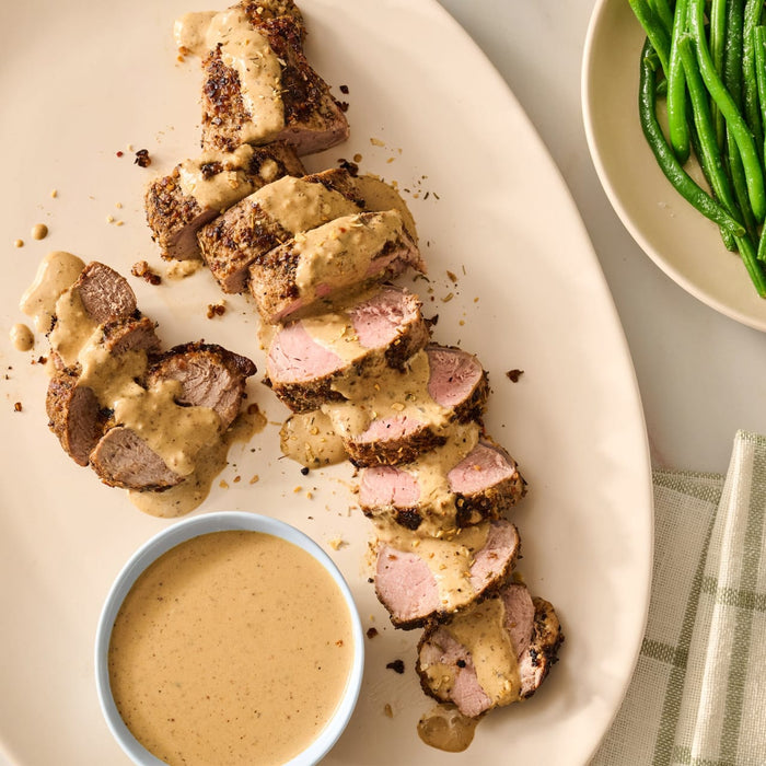 A sliced pork tenderloin is drizzled with a creamy sauce and garnished with Mccormick Onion And Herb Seasoning Crystal 4.16 Oz alongside a bowl of extra sauce and fresh green beans.