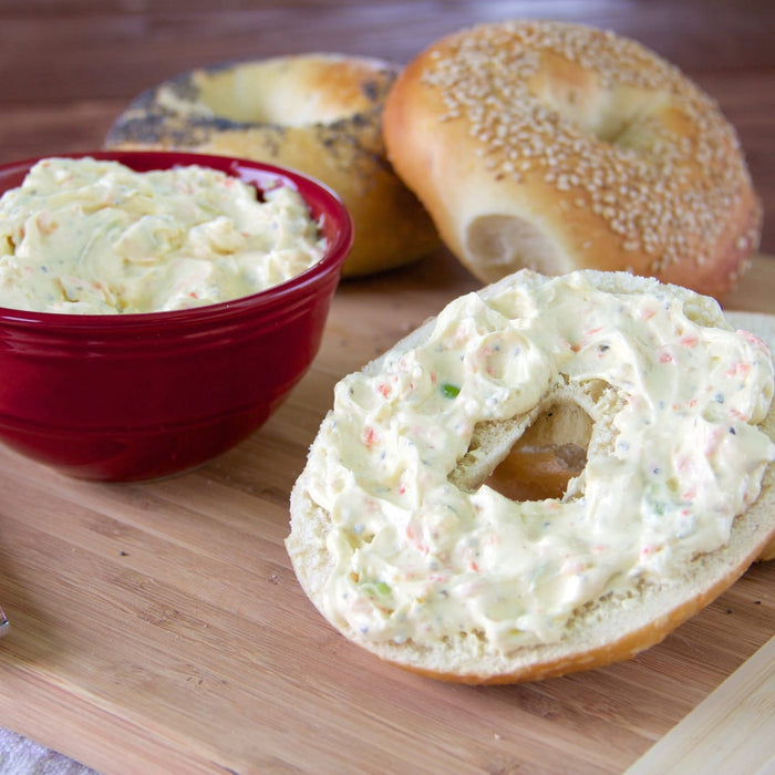 A bagel with cream cheese and a bowl of cream cheese sit on a wooden cutting board.