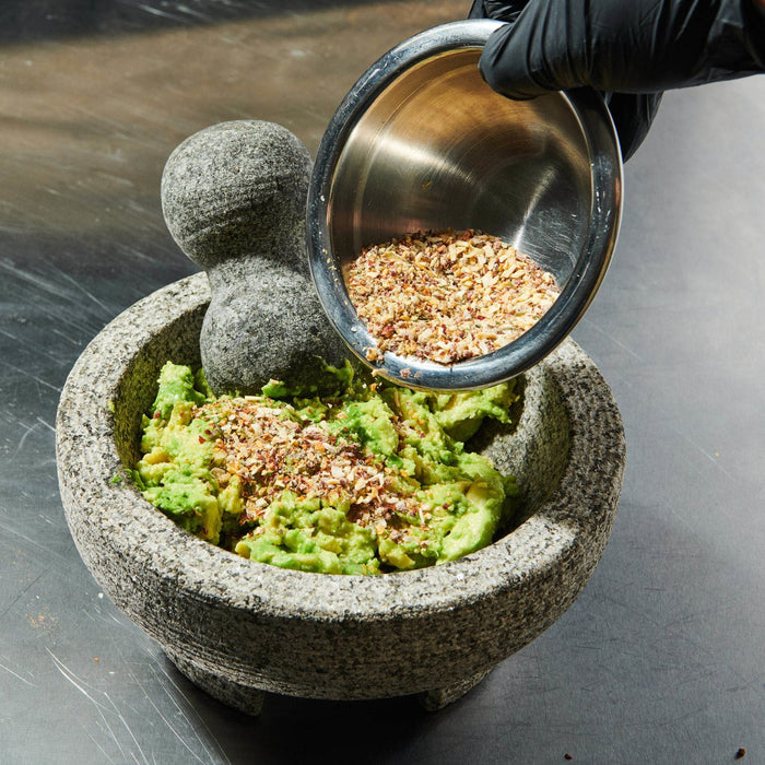 ['A hand in a black glove pours seasoning onto mashed avocado in a stone mortar and pestle.']