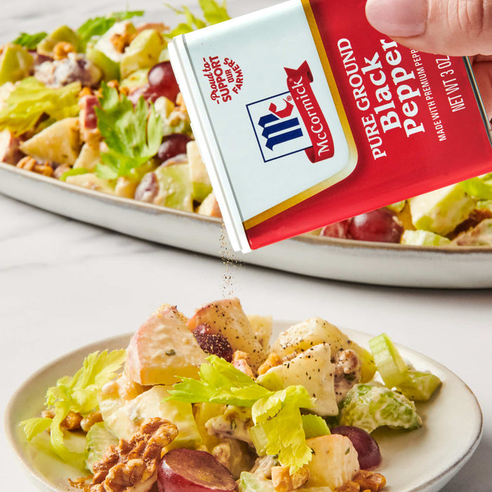 ['A hand sprinkles McCormick Pure Ground Black Pepper on a Waldorf salad.']
