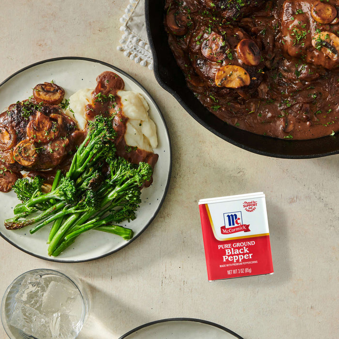['McCormick Pure Ground Black Pepper next to a plate of beef patties with mashed potatoes and broccoli and a skillet of beef patties with mushroom gravy.']