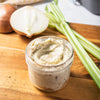 ['A jar of seasoned butter sits on a wooden cutting board with celery and onions.']