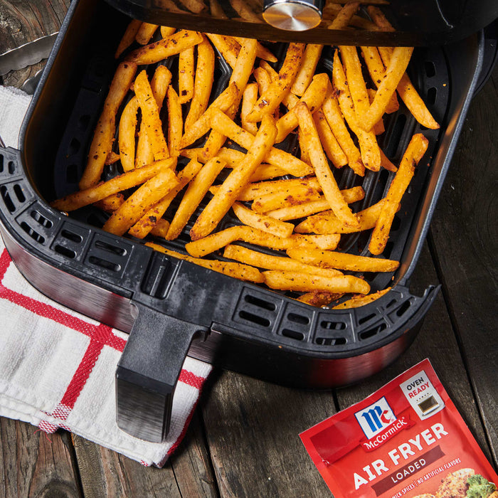 ['A close-up of a black air fryer with crispy french fries inside, next to a McCormick Air Fryer Loaded seasoning packet.']