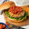 ['A hamburger with tomato salsa on a bun with lettuce and sweet potato fries on a wooden cutting board.']