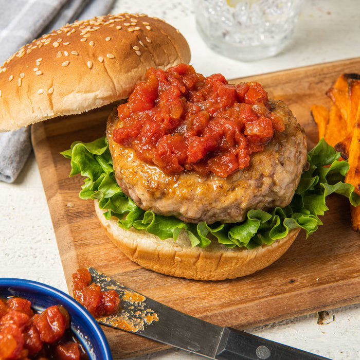 ['A hamburger with tomato salsa on a bun with lettuce and sweet potato fries on a wooden cutting board.']