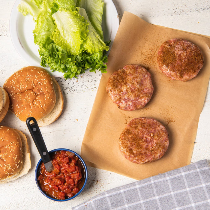 ['A white table with burger patties, buns, lettuce, tomato salsa, and a checkered napkin.']