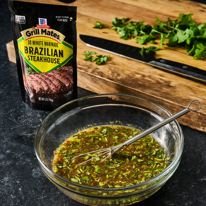 A package of McCormick Grill Mates Brazilian Steakhouse 30 Minute Marinade is next to a bowl of marinade with a whisk a knife and cilantro on a cutting board.