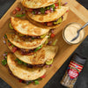 A wooden board displays stacked Jack's Famous Smash Tacos with a side of dipping sauce and a bottle of Jack's Famous Smash seasoning.