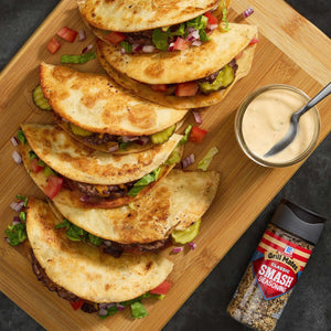 A wooden board displays stacked Jack's Famous Smash Tacos with a side of dipping sauce and a bottle of Jack's Famous Smash seasoning.