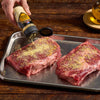 Two raw steaks are being seasoned with McCormick Grill Mates Montreal Steak Seasoning on a metal tray.