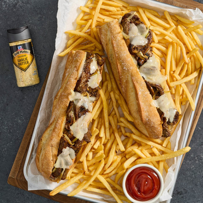 Two cheesesteak sandwiches with fries and a bottle of McCormick Montreal Steak Seasoning.