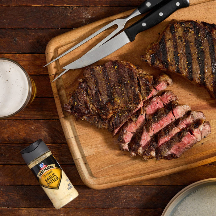 A grilled steak, sliced and served on a wooden cutting board with a carving fork and McCormick Grill Mates Montreal Steak Seasoning, alongside a glass of beer.