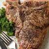 ['A close-up of a perfectly cooked T-bone steak with parsley and a fork and knife.']