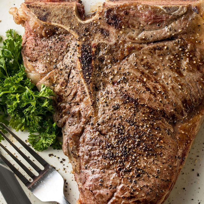 ['A close-up of a perfectly cooked T-bone steak with parsley and a fork and knife.']