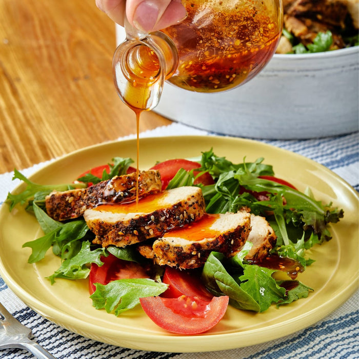 A hand pours a honey sesame dressing over a salad with grilled chicken and tomatoes.