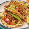 ['Three tacos filled with meat, lettuce, tomato, avocado, and onion on a white plate.']