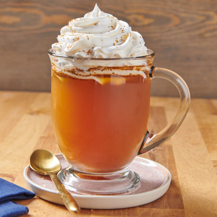 A glass mug filled with a warm beverage topped with whipped cream sits on a saucer with a spoon.
