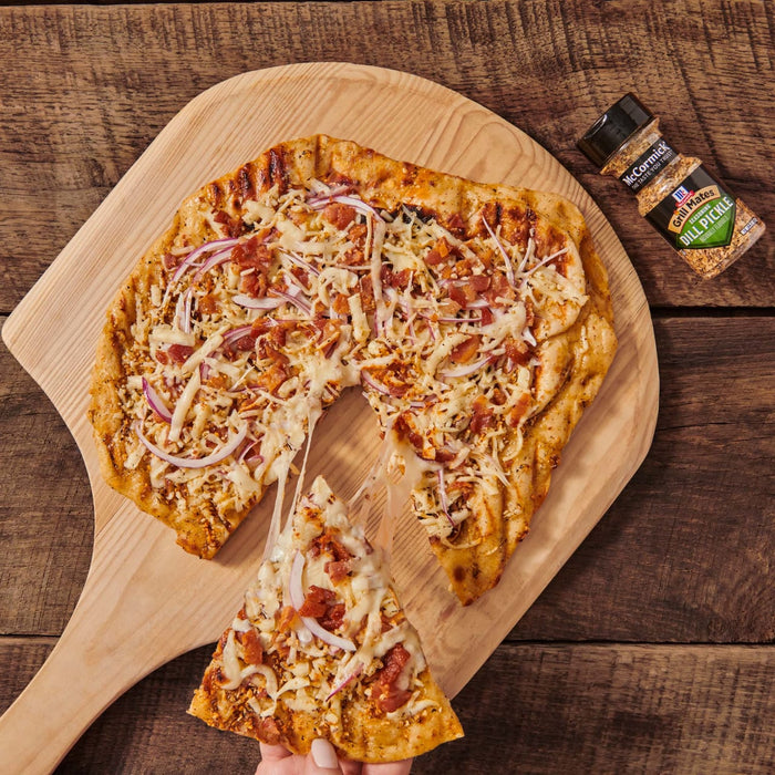 ['A pizza topped with bacon, cheese, and red onion on a wooden cutting board with a jar of McCormick Grill Mates Dill Pickle seasoning.']