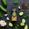 A bottle of Kernel Season's Dill Pickle seasoning is surrounded by cucumbers, garlic, dill, and peppercorns.