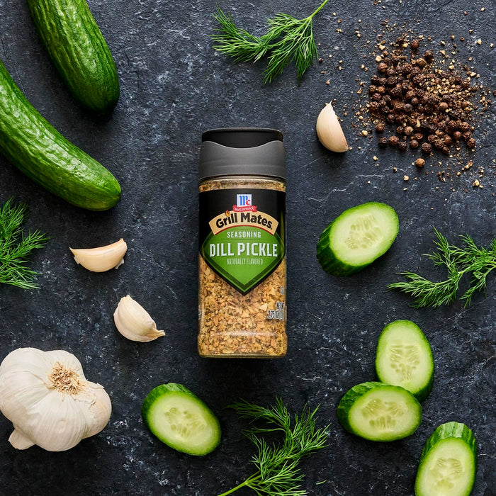 A bottle of Kernel Season's Dill Pickle seasoning is surrounded by cucumbers, garlic, dill, and peppercorns.