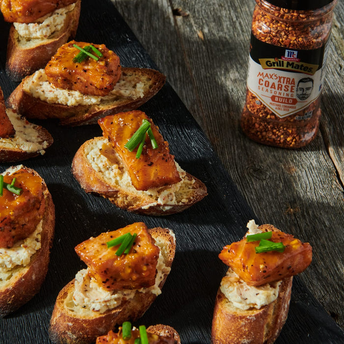 ["A close-up of a black slate board with six crostini topped with a creamy spread and a piece of grilled salmon, with a bottle of McCormick Grill Mates Max's Extra Coarse Seasoning in the background."]