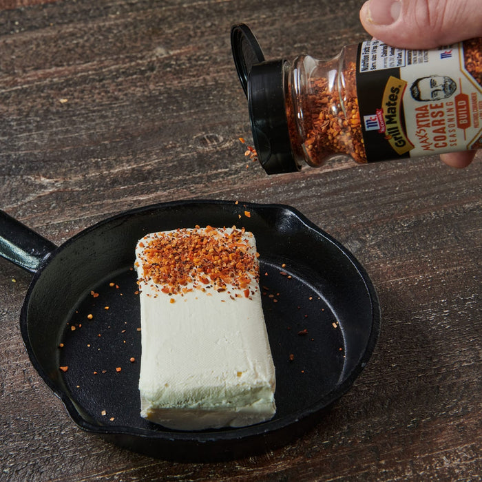 ["A hand sprinkles McCormick Grill Mates Max's Extra Coarse Seasoning Build on a block of cream cheese in a cast iron skillet."]
