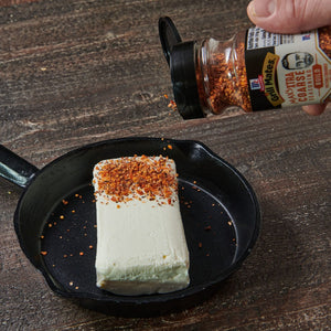 ["A hand sprinkles McCormick Grill Mates Max's Extra Coarse Seasoning Build on a block of cream cheese in a cast iron skillet."]